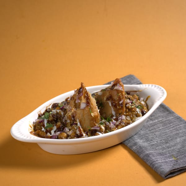 Two samosas topped with sauces, chopped onions, and chickpeas in a white dish.