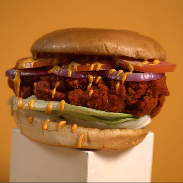 Crispy red patty burger with lettuce, tomato, red onion, and yellow sauce on a bun.